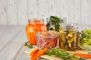 Barattoli di vetro contenenti diverse verdure fermentate, come carote, cetrioli e cavoli, su un tavolo di legno chiaro, illustrando alcune ricette tipiche della cucina fermentata