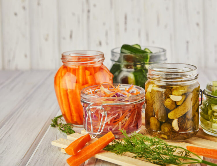 Barattoli di vetro contenenti diverse verdure fermentate, come carote, cetrioli e cavoli, su un tavolo di legno chiaro, illustrando alcune ricette tipiche della cucina fermentata