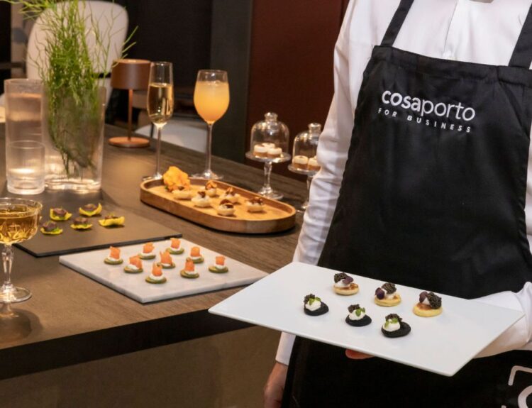 Un cameriere con grembiule "Cosaporto for Business" presenta un vassoio di finger food gourmet, parte del servizio "Cosaporto Digital Catering", circondato da eleganti drink e antipasti su un tavolo raffinato.